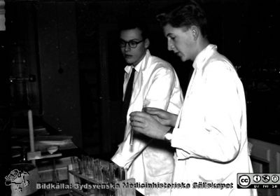 Kurslaboration på institutionen för medicinsk kemi i Lund 1957
Från vänster ej identifierad amanuens och Björn Östberg vid laboratoriebänken. Foto och bildkälla Sven Åke Hedström.
Nyckelord: Institution;Medicinsk kemi;Lund;Kurslaboration