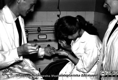 Kurslaboration på institutionen för medicinsk kemi i Lund ht 1957
Kerstin Håkansson (senare gift  Lilja) sticker Berndt Ehinger med en kapillärblodslancett för att denne sedan skall kunna ta ett kapillärblodprov med den blodprovspipett han håller i handen. Åskådaren är Göran Eklundh. Foto och bildkälla Sven Åke Hedström.
Nyckelord: Kurslaboration;Medicinsk kemi;Institution;Lund;Fakultet