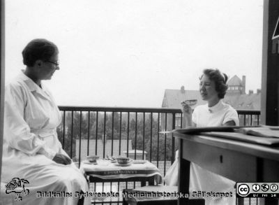 Kaffepaus på en balkong högt upp på norrfasaden på Jubileumskliniken i Lund. 
Folkskoleseminariet i fonden. Till vänster på balkongen kanske en patient och till höger en SSSH-sjuksköterska. Foto före 1957 då panncentralen byggdes bortom seminariet  med hög skorsten.
Nyckelord: Jubileumsklinik;Sjuksköterska;Kaffepaus