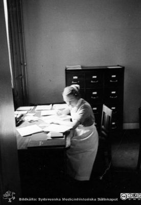 En SSSH-sjuksköterska  i journalarkivet på radiologiska kliniken i Lund. 
Ett måttligt stort journalskåp i bakgrunden. Foto kanske på 1950-talet. Från Syster Elinas fotoalbum (Elina Holmberg?) .
Nyckelord: Radiologisk;Onkologisk;Jubileumsklinik;Lasarettet i Lund;Journalföring