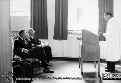 Kungligt besök på radiologiska kliniken i Lund 1952. 
Kung Gustaf VI Adolf näst längst bort på första stolsraden med en galonerad adjutant i sjöofficersuniform på sin vänstra sida. Möjligen är det amiral Stig H:son Ericsson. Kiinikchefen docent Bertil Ebenius står i talarstolen. Från Syster Elinas fotoalbum (Elina Holmberg?).
Nyckelord: Radiologisk;Onkologisk;Jubileumsklinik