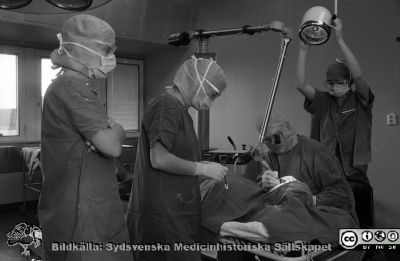 Laseroperation 1981
Sjukhusfotograferna i Lund. Pärm Negativ 1981:1. 81-11-13. Laseroperation på urologiska kliniken. Instrumentet har stel spegelljusledare, som de tidiga långvågiga koldioxidlasrarna hade. Kanske är operatören docent Eric Lindstedt. Rimligen är lasern ny eftersom en professionell fotograf har tagit bilder på operationssalen. 
Operatören provar att använda den på något i patientens ansikte, ett ovanligt ställe för en urolog. 
Han bär mössa och handskar, men inte munskydd. De assisterande bär handskar, munskydd och mössor, men är inte sterilklädda. Från negativ
Nyckelord: Lasarettet;Lund;Universitetssjukhuset;USiL;Kirurgisk;Klinik;Operation;Laser