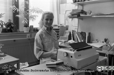 En okänd men mycket typisk medicinsk sekreterare 1984
Hon har en tidstypisk elektrisk skrivmaskin av märket Facit, en lika tidstypisk dsdiktafon för uppspelning av kassettband, styrd av en fotpedal och i bakgrunden en snabbtelefon.
Nyckelord: Lasarettet;Lund;Universitetssjukhuset;USiL;Administration