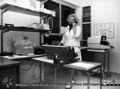 Receptionisten fru Brita Jäderström på infektionskliniken i Lund.
Foto ur ett album tillägnat professor Karl Emil Thulin på hans 60-årsdag, 1975. Sekreterare / receptionist fru Brita Jäderström  i infektionsklinikens reception, klinikens vänlighet utåt. Notera den gamla tunga telefonen i bakelit, den stora skrivmaskinen och den ännu större maskinen vid bortväggen, troligen för prägling av patientbrickor.

Nyckelord: Lasarettet;Lund;Universitetssjukhuset;USiL;Infektion;Epidemiologisk;Kliniken