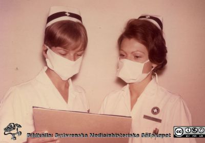 Till höger sjuksköterskan Hanna-Kerstin Einarsson enligt namnbrickan.
Karl Emil Thulins efterlämnade fotoalbum från 1970-talet, i infektionsklinikens bibliotek.Två sjuksköterskor på infektionskliniken i munskydd.  Från originalfoto
Nyckelord: Lasarettet;Lund;Universitetssjukhuset;USiL;Infektion;Klinisk;Epidemiologisk
