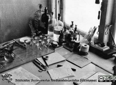 Ett arbetsbord på laboratoriet på hudkliniken i Lund från mitten av 1900-talet
Ur yrkesdermatologen Sigfrid Fregerts bildsamling från hud- och könskliniken i Lund och från yrkesdermatologiska avdelningen i Lund. Ett kliniskt laboratorium på hud- och könskliniken. Spetsglasen användes för att blanda till Neosalvarsanlösning för injektion. Preparatet hade fortfarande viss användning som behandling mot syfilis. Gammalt foto, kanske från 1950-talet innan penicillin ersatte Salvarsanet och senast 1972 då kliniken fick nya lokaler. Foto.
Nyckelord: Lasarettet;Lund;Universitetssjukhuset;USiL;Hud- och könsklinik;Dermatologisk;Venerologisk;Avdelning