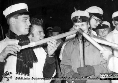 Lucia 13/12 1956 
? Valter Andersson, kr. Frank Werneman, Gunilla Nilsson. Tågets facklor tänds på Universitetsplatsen utanför Akademiska Föreningen i Lundagård.
Nyckelord: Luciafest;Luciatåg;Västsvenska nationerna;Lundagård;Universitetsplatsen