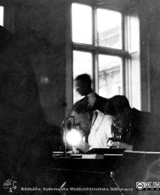 1928 års histologikurs i Lund
Fotoalbum märkt Histologikursen anno 1928. 
Detta album innehåller bilder från dåvarande histologiska avdelningen på anatomiska institutionen i Lund på Biskopsgatan 7. Liksom de övriga i serien skapades det av kursdeltagarna och tillägnades kursledaren, professor Torsten Hellman, som uppskattning av hans lärargärning. Bilden är inte namnsatt. Den visar professor Torsten Hellman i ljus laboratorierock vid ett kursmikroskop tillsammans med medicine studeranden i avdelningens mikroskoperingssal.
Bildkälla Stiftelsen Locus Medicus Lundensis 250317.
Nyckelord: Locus Medicus Lundensis;Medicinska fakulteten i Lund;Histologiska avdelningen;Sydsvenska Medicinhistoriska Sällskapet;SMHS;Anatomiska Institutionen;Studenter;Lärare