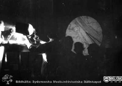 1928 års kurs i histologi i Lund
Fotoalbum märkt Histologikursen anno 1928. 
Detta album innehåller bilder från dåvarande histologiska avdelningen på anatomiska institutionen i Lund på Biskopsgatan 7. Liksom de övriga i serien skapades det av kursdeltagarna och tillägnades kursledaren, professor Torsten Hellman, som uppskattning av hans lärargärning. Bilden visar en föreläsning där en mikroskopisk bild projiceras på väggen med hjälp av ett tidigt projektionsmikroskop, kanske byggt på institutionen. Kommersiellt byggda projektionsmikroskop fanns visserligen från firma Zeiss i Tyskland, men de såg inte ut som på bilden. Ingen bildtext.  
Bildkälla Stiftelsen Locus Medicus Lundensis 250317.
Nyckelord: Locus Medicus Lundensis;Medicinska fakulteten i Lund;Histologiska avdelningen;Sydsvenska Medicinhistoriska Sällskapet;SMHS;Anatomiska Institutionen;Studenter;Lärare