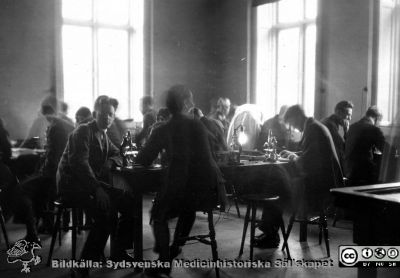 1928 års kurs i histologi i Lund
Fotoalbum märkt Histologikursen anno 1928. 
Detta album innehåller bilder från dåvarande histologiska avdelningen på anatomiska institutionen i Lund på Biskopsgatan 7. Liksom de övriga i serien skapades det av kursdeltagarna och tillägnades kursledaren professor Torsten Hellman som uppskattning av hans lärargärning. Bildtext  saknas. Bilden visar rimligen studenter och en amanuens i ljus rock på mikroskoperingssalen. Flera har rört på sid under den något långa exponeringen.
Bildkälla Stiftelsen Locus Medicus Lundensis 250317.
Nyckelord: Locus Medicus Lundensis;Medicinska fakulteten i Lund;Histologiska avdelningen;Sydsvenska Medicinhistoriska Sällskapet;SMHS;Anatomiska Institutionen;Studenter;Lärare