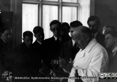 Fotoalbum  "Kursen i histologi V. T. 1933.
Fotoalbum  "Kursen i histologi V. T. 1933. Bildtext: "Chefen docerar". Professor Torsten Hellman berättar vid ett mikroskop. Medicine studerande i histologiska avdelningens mkroskoperingssal på anatomiska institutionen i Lund. 
Bildkälla stiftelsen Locus Medicus Lundensis och Sydsvenska Medicinhistoriska Sällskapet / SMHS. Fotograf okänd.
Nyckelord: Locus Medicus Lundensis;Medicinska fakulteten i Lund;Histologiska avdelningen;Medicinska fakulteten i Lund;Sydsvenska Medicinhistoriska Sällskapet;Anatomiska Institutionen;Student